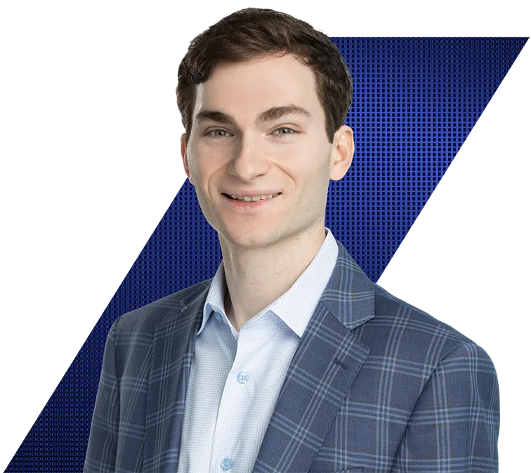 Headshot of a smiling young man with brown hair and green eyes, wearing a blue plaid suit jacket and light blue shirt, against a black background with a blue geometric pattern.