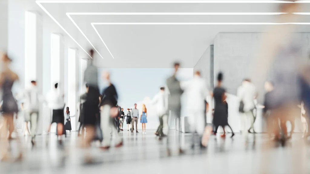 Blurred image of a busy, modern hall with people moving. White ceiling with geometric linear lights, reflective floor.