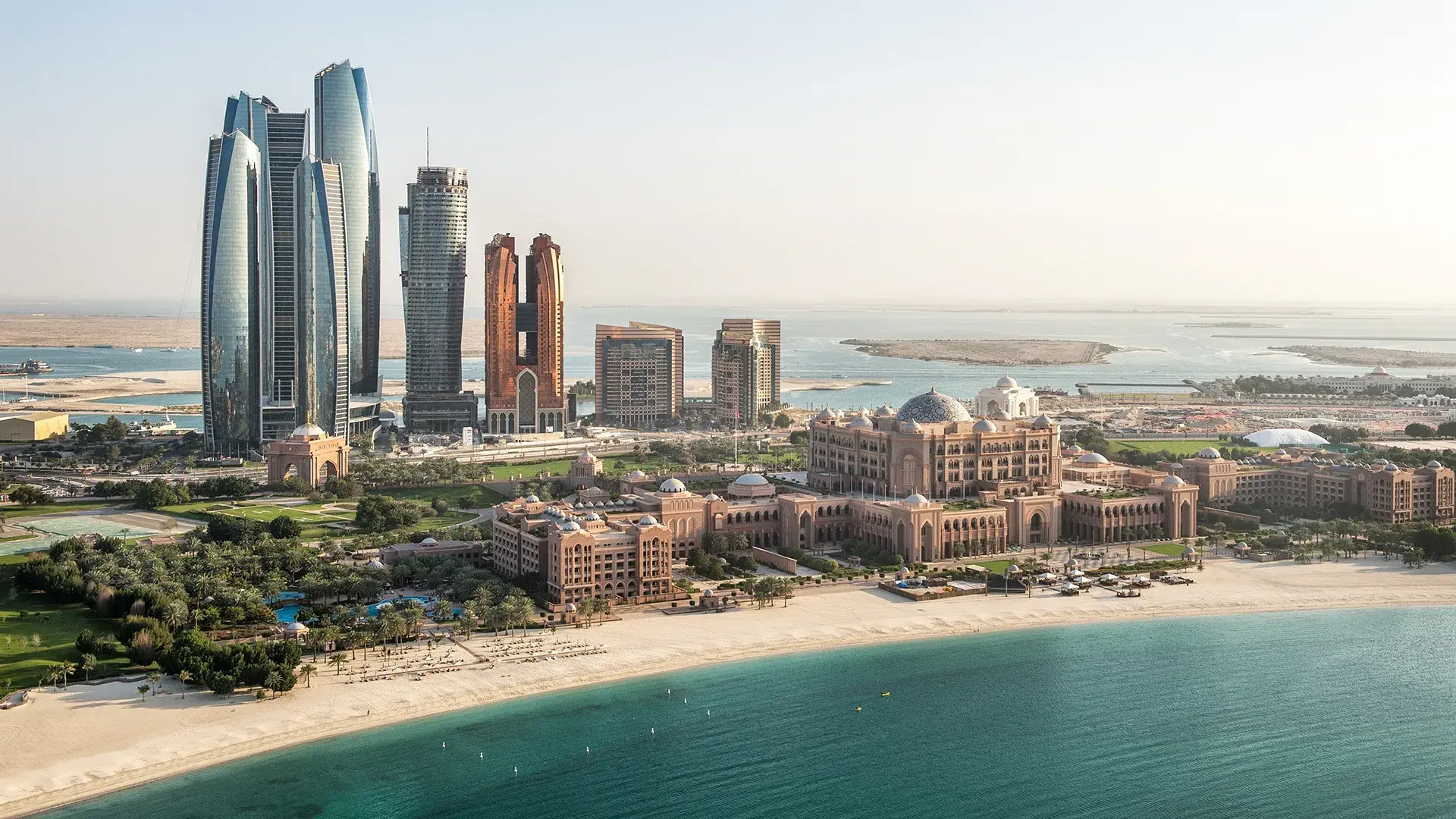 Aerial view of Abu Dhabi showing the grand Emirates Palace hotel, the sleek Etihad Towers skyscrapers, a pristine sandy beach, and turquoise ocean water.