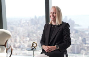 A smiling Ashley MacNeill in a black blazer sits in front of a large window with a blurred city skyline view.