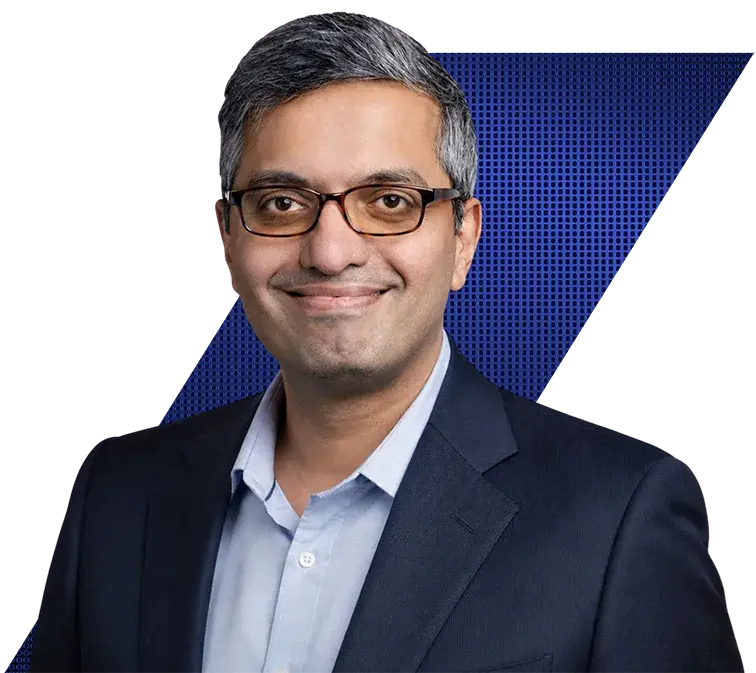 Headshot of a smiling man with graying hair and glasses, wearing a blue shirt and dark suit jacket. The background is black with a blue dotted geometric pattern.