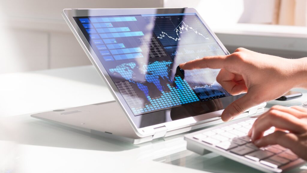 A person's hand touches a laptop screen displaying a data dashboard with graphs, charts, and a world map, while another hand types on a separate keyboard.