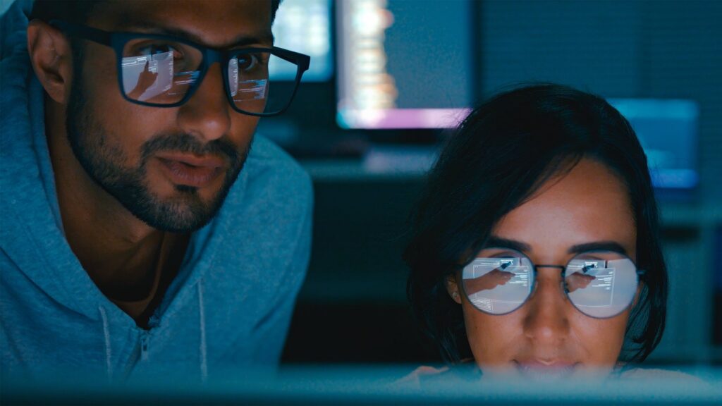Two people, a man and a woman, wearing glasses, looking at a screen. Reflections of computer code and a hand pointing are visible in their glasses.