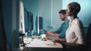 A woman with short curly hair and glasses, wearing headphones, works at a computer in a dim office. A man works in the blurred background.