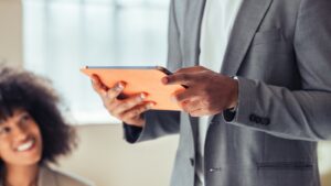 A person in a grey suit holds an orange-cased tablet. Another person with curly hair smiles in the blurred background.