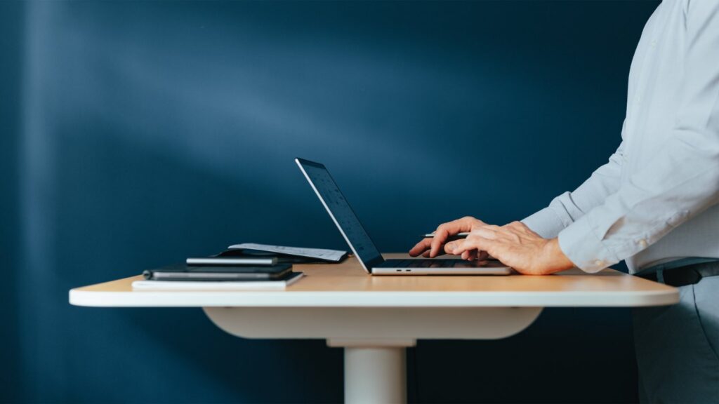 A person in a light blue shirt stands at a light wooden desk, typing on a silver laptop with a graph on its screen. A notebook, pen, and papers are beside the laptop. The background is a dark blue wall.