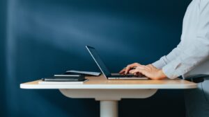 A person in a light blue shirt stands at a light wooden desk, typing on a silver laptop with a graph on its screen. A notebook, pen, and papers are beside the laptop. The background is a dark blue wall.